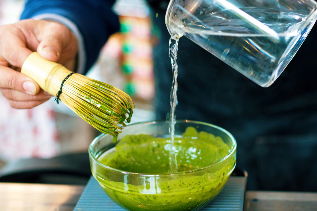 Pouring hot water into a bowl of ceremonial grade matcha tea while holding a bamboo whisk (chasen), demonstrating the traditional matcha preparation process